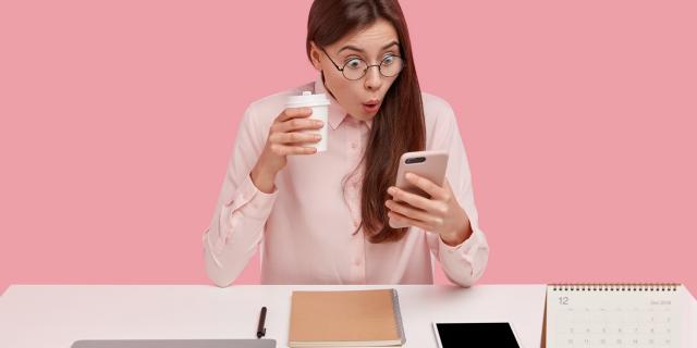 Photo of attractive young woman reads shocking news on mobile phone, watches video in social networks, drinks coffee from disposable cup, being office perfectionist, uses electronic gadgets.