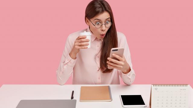 Photo of attractive young woman reads shocking news on mobile phone, watches video in social networks, drinks coffee from disposable cup, being office perfectionist, uses electronic gadgets.